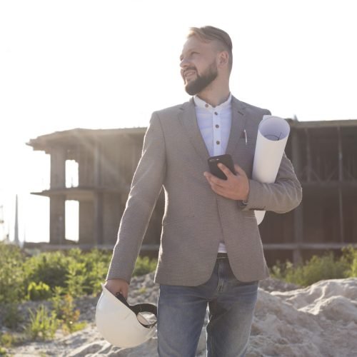 portrait-smiling-architect-man-holding-cellphone-hard-hat-with-blueprint-construction-site